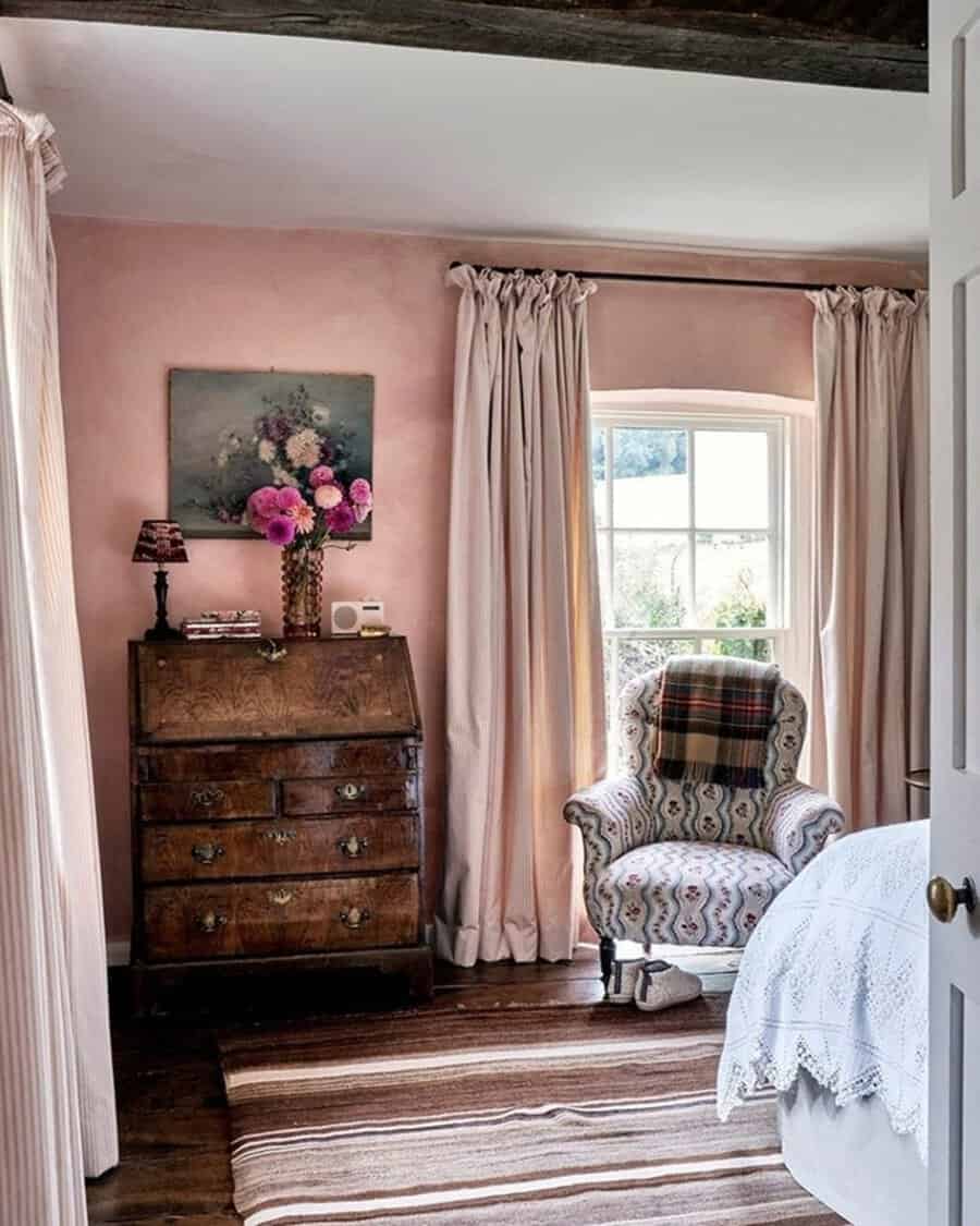 Vintage bedroom with soft pink walls, antique wooden furniture, flowing curtains, and a cozy armchair for a warm, inviting atmosphere.