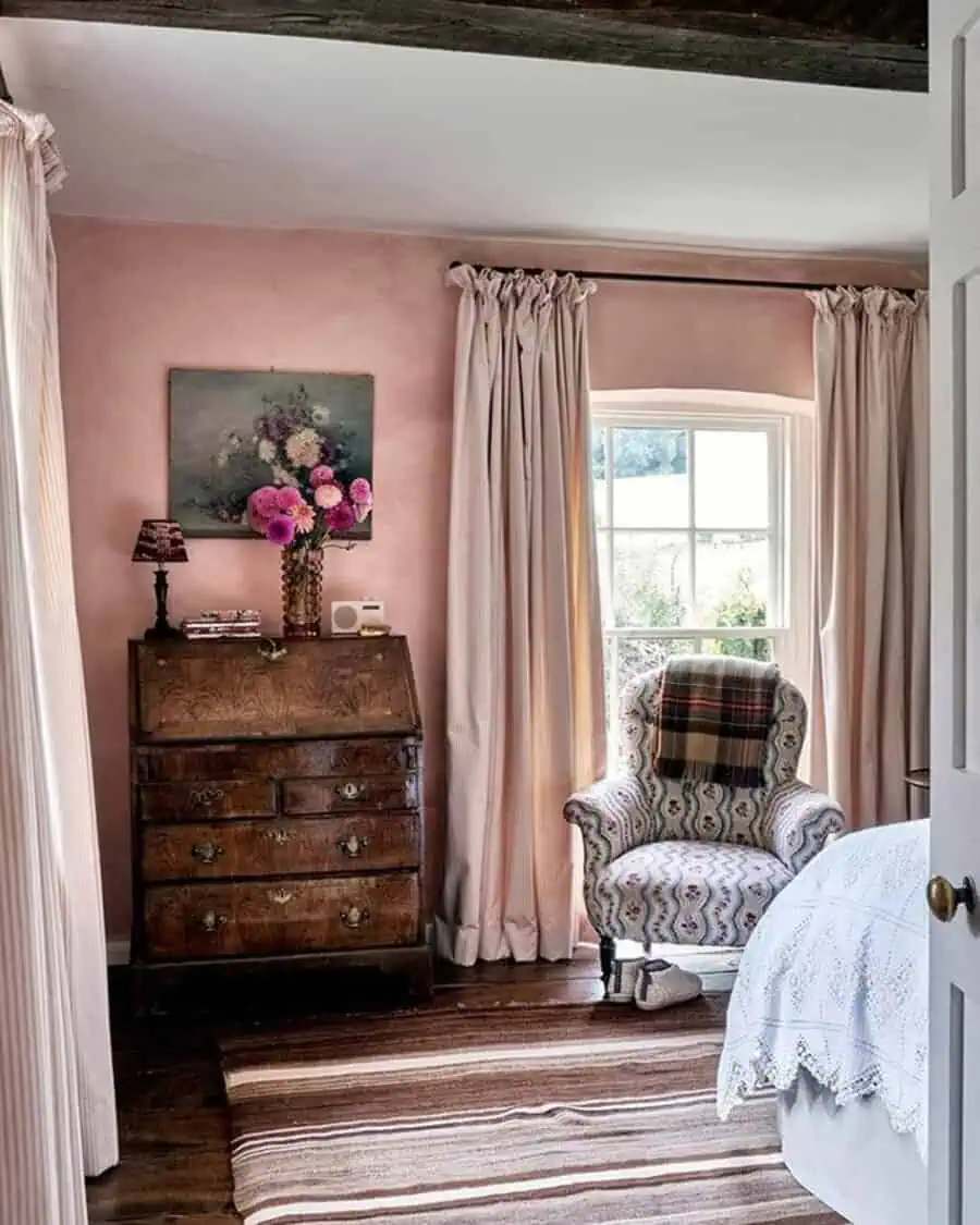 Vintage bedroom with soft pink walls, antique wooden furniture, flowing curtains, and a cozy armchair for a warm, inviting atmosphere.