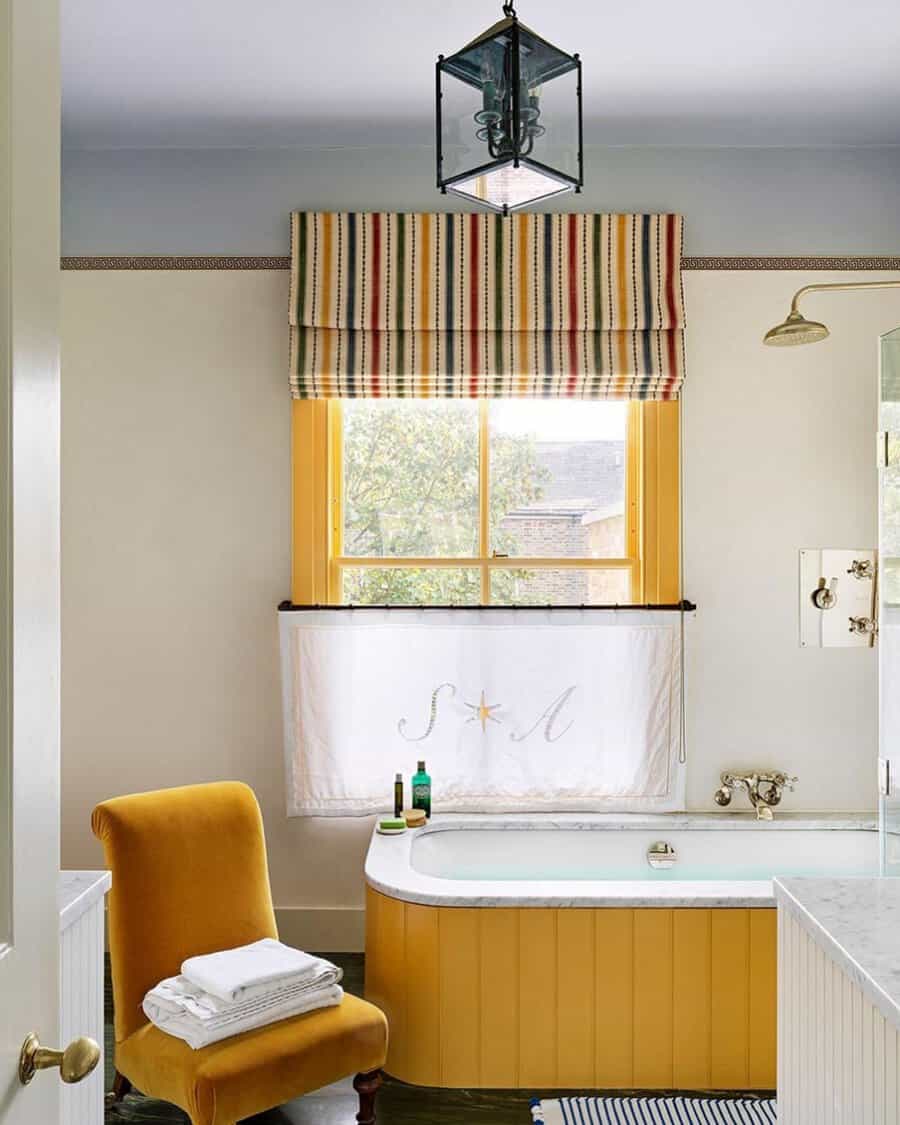 Bathroom with a yellow bathtub, striped window shades, vintage chair, and marble accents for a warm, classic look.