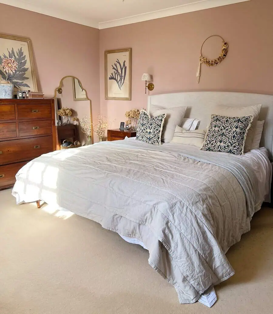 Muted pink walls with framed botanical prints and a dried flower wreath create a warm, elegant backdrop for a cozy bedroom.