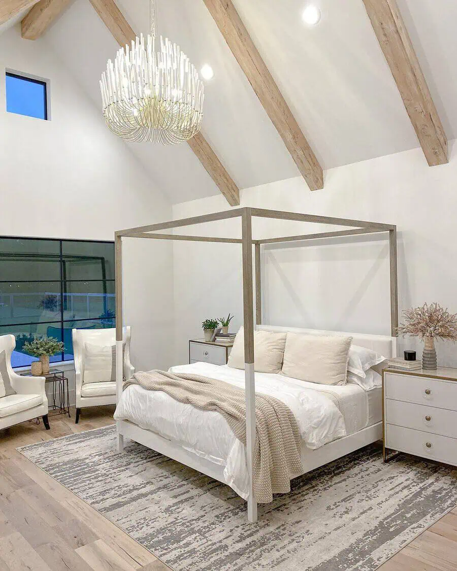 Bright bedroom with tall wood beams, a large white chandelier, four-poster bed, and neutral decor creating a cozy, airy atmosphere.