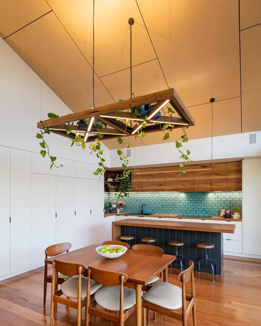 Open kitchen with a wood panel vaulted ceiling, teal subway tile, and a hanging greenery light fixture above a wooden dining set.