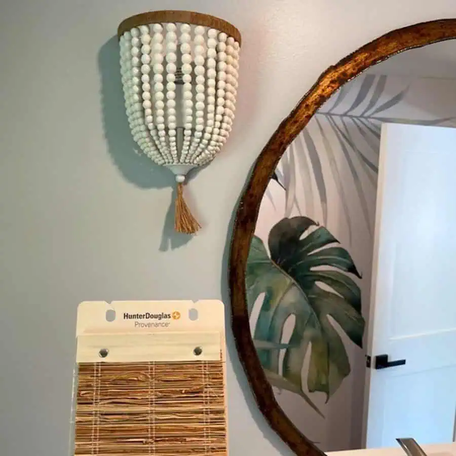 Large wooden-framed mirror above a modern vanity, with greenery visible through the window and soft lighting from vintage sconces in a bathroom.